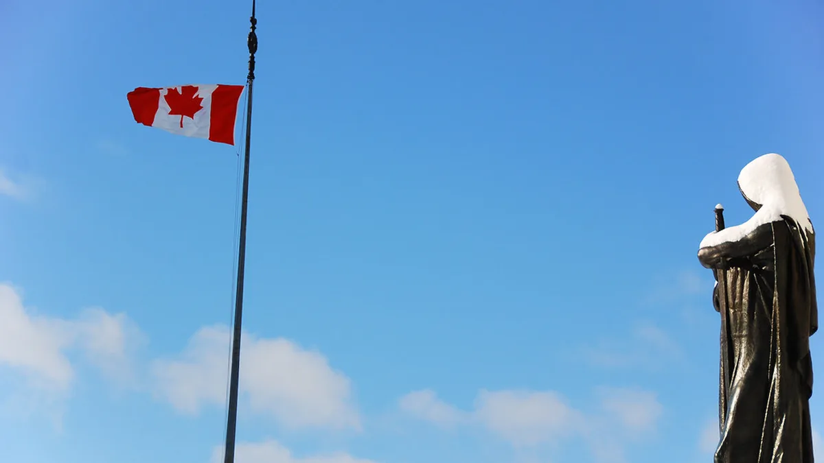Canadian flag flying against a blue sky next to a bronze statue